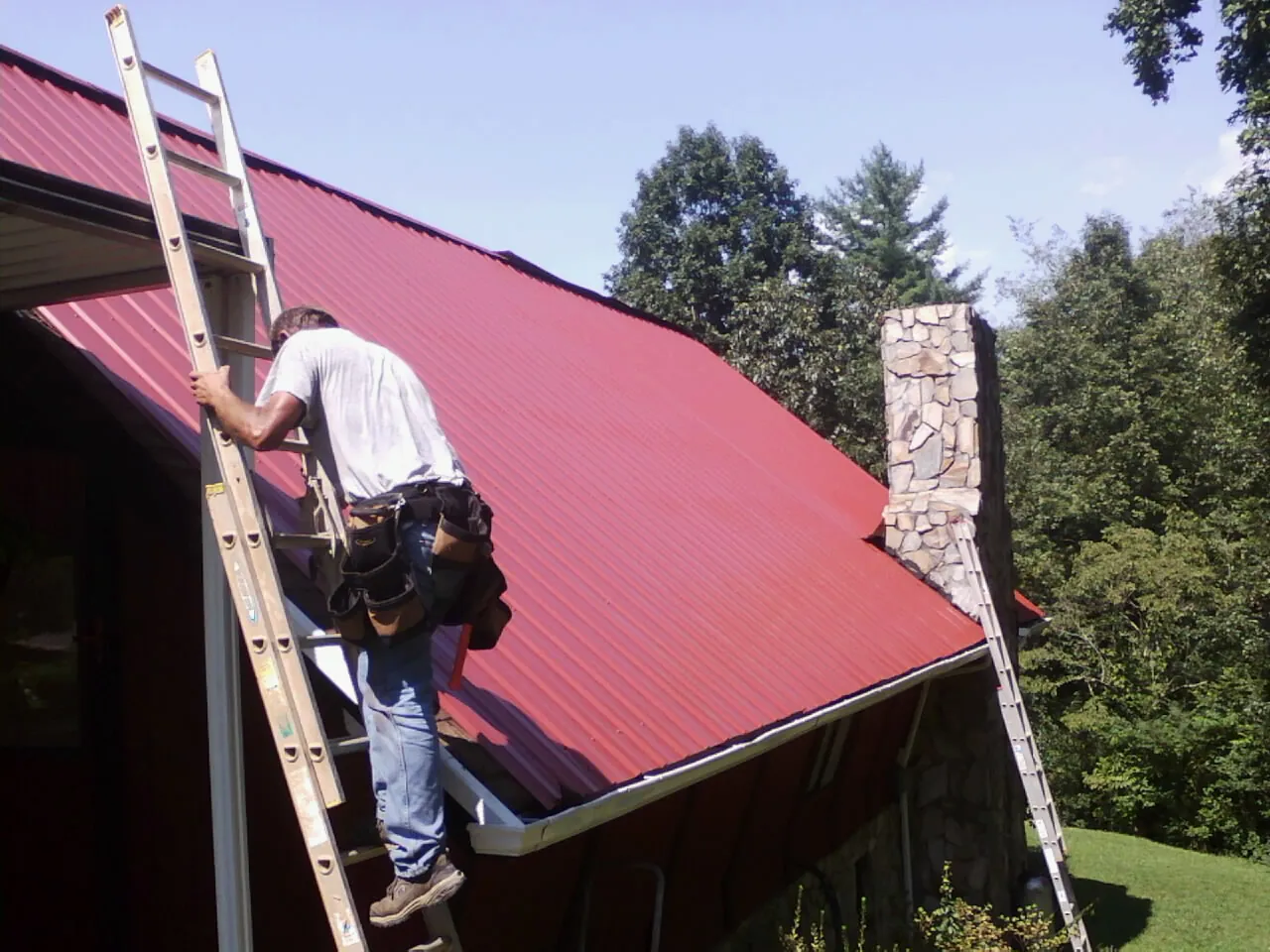 Roofer on a ladder beside a red metal roof and stone chimney — Soffit & Fascia Repair by Roofing Brothers in Hewitt